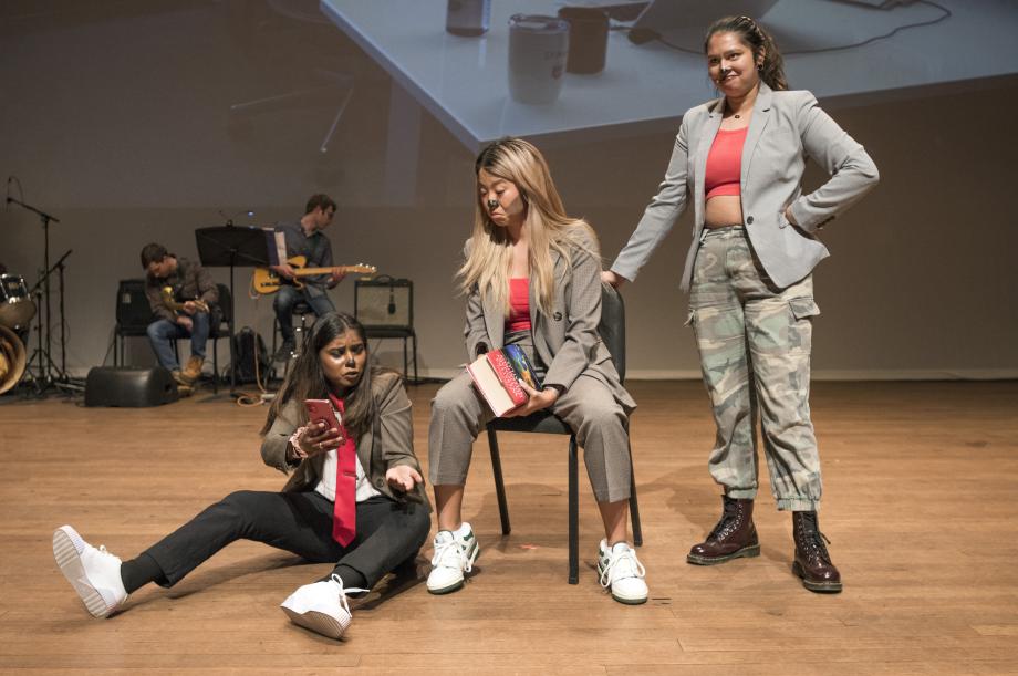 Three students on stage, one sitting on the floor, one sitting in a chair, and one standing. 