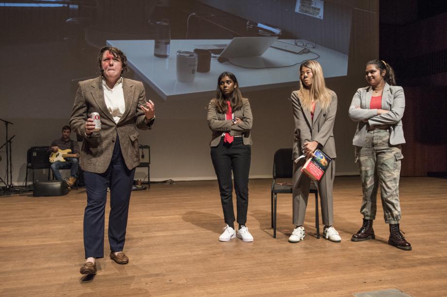 Kelly Gregg stands on stage with three students portraying hyenas stand in a row behind her.