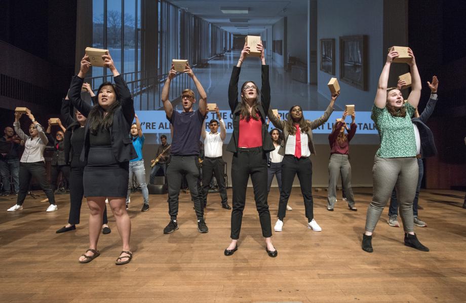 Members of the cast sing while holding brown takeout boxes above their heads.