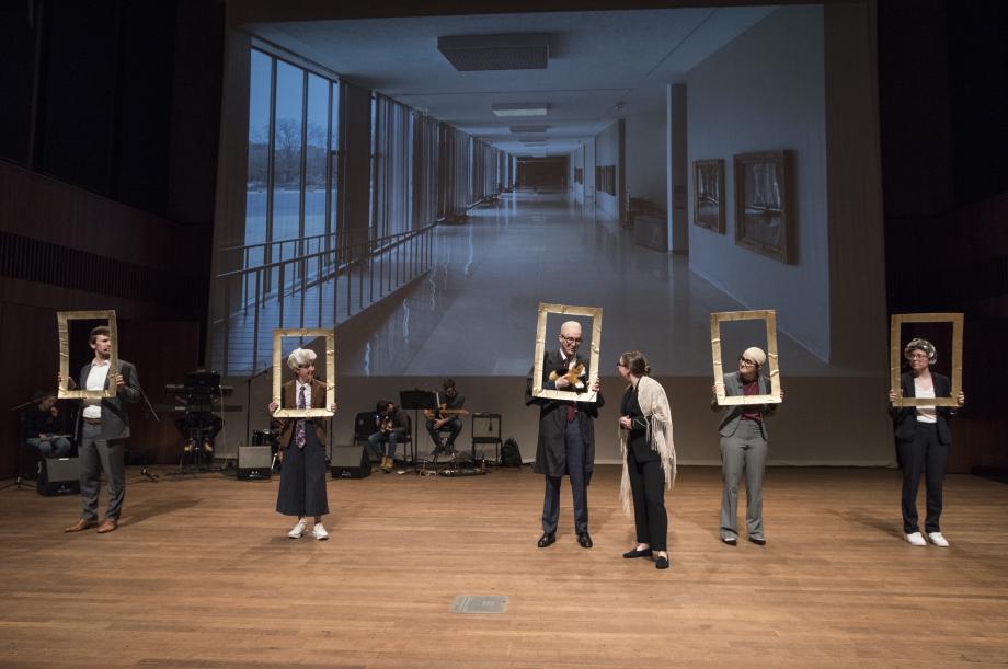 Students holding portrait frames around their faces line up before a screen showing a photo of the classroom hallway.