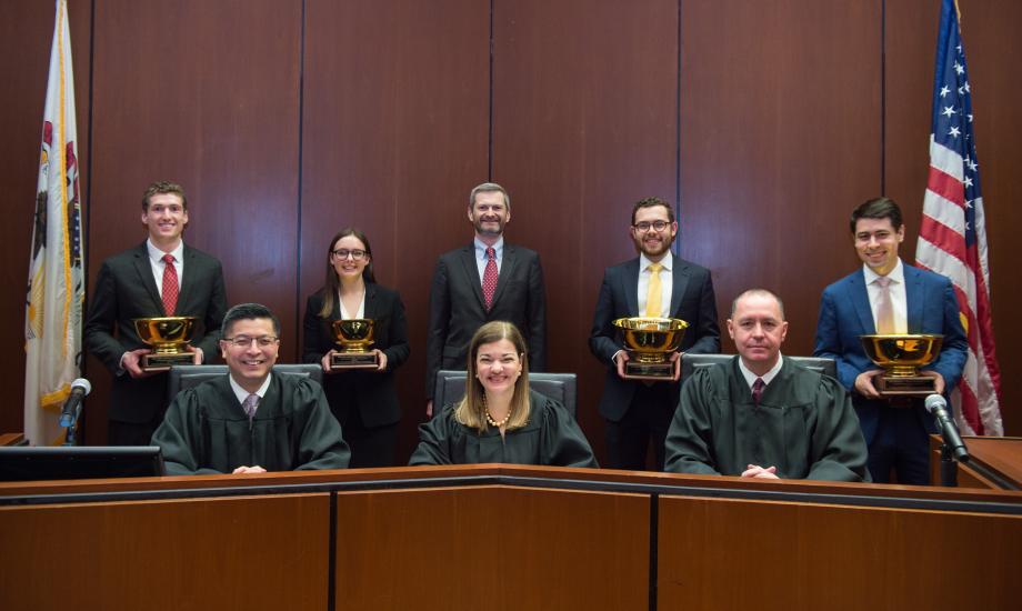 Four students holding their trophies pose behind the three judges at the bench.