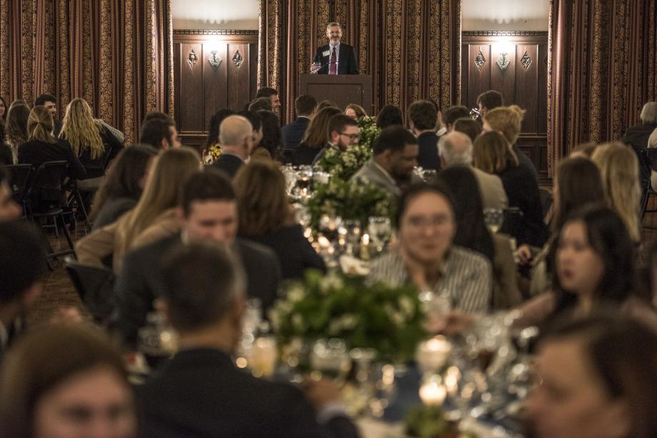 Dean Miles speaks from the podium with faculty, students, and staff sitting at tables in the foreground