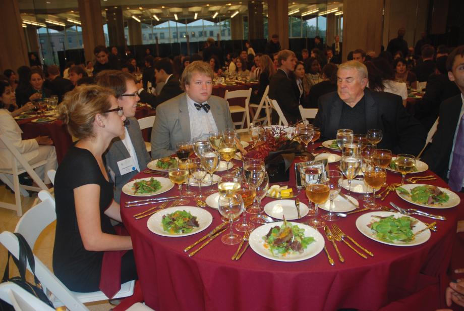 A dinner table with red cloth and with salad-like food out. Hutchinson sits there and talks to students.