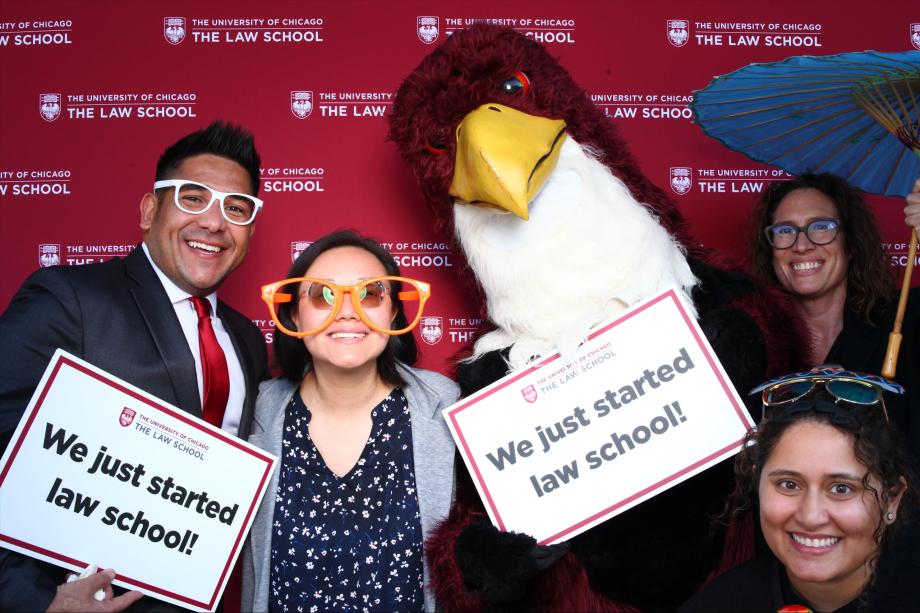 Four people and mascot pose in front of a branded backdrop holding signs, "we just started law school!"