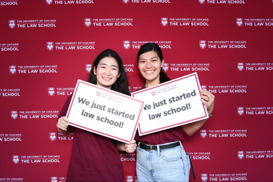 Two people pose in front of a branded backdrop holding signs, "we just started law school!"