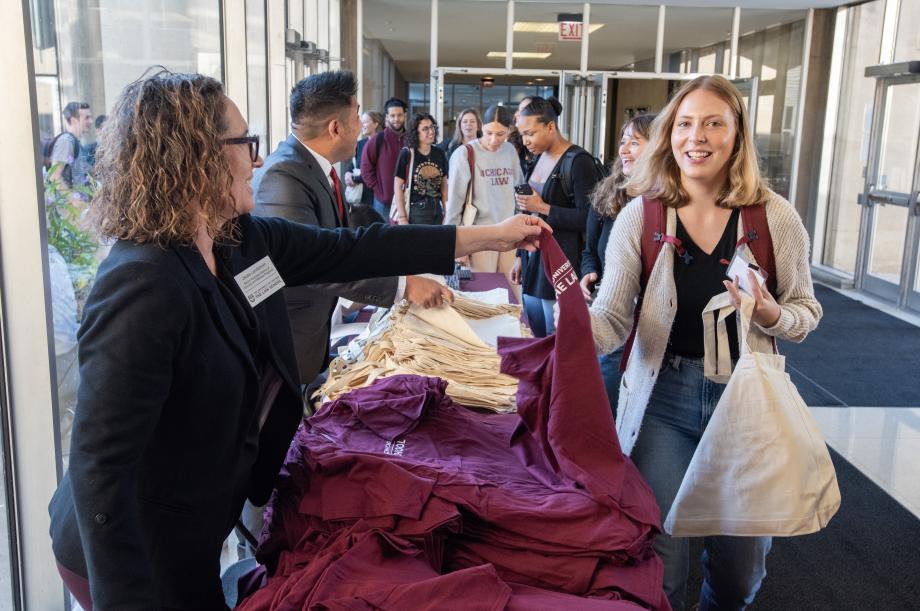 On one side of a table, staff members hand out t-shirts and tote bags to a line of students.
