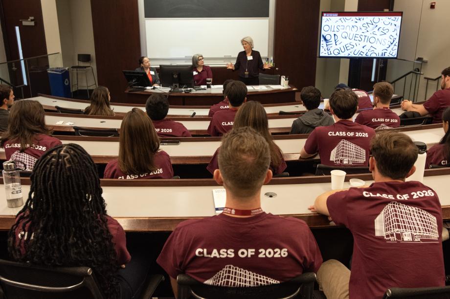 Staff members present from the front of the classroom with students sitting in rows in the foreground.