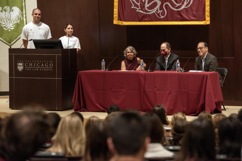 Three faculty members sit behind a panel with two orientation leaders standing at the podium.