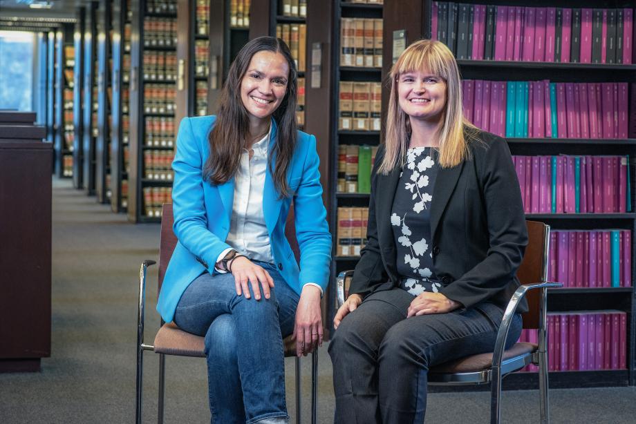 Two women sit side by side with library book stacks behind them.