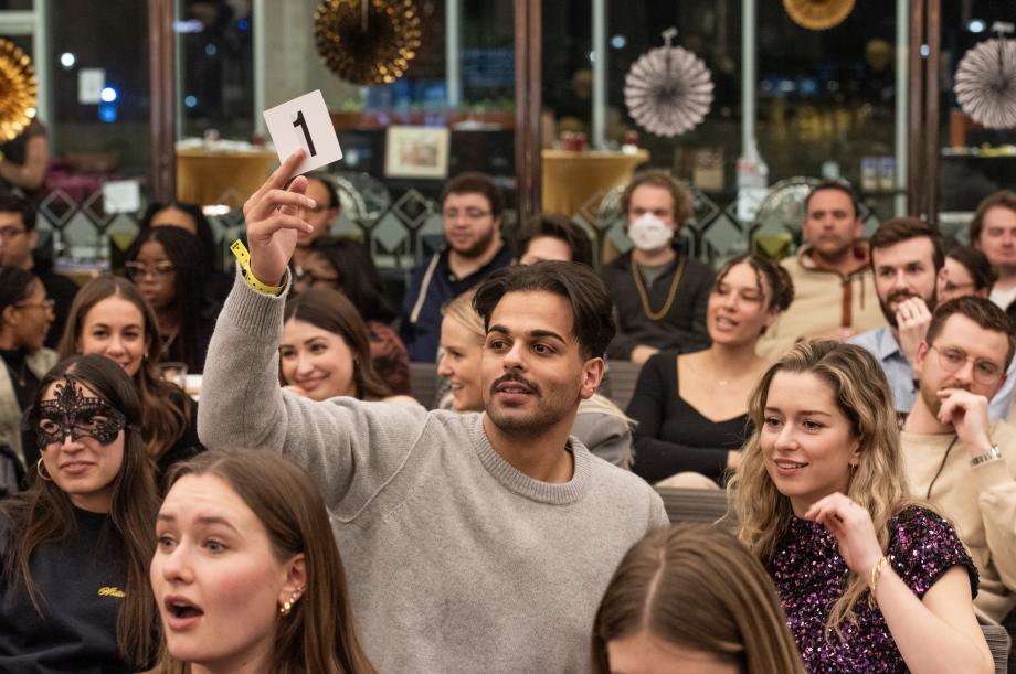 A man in a grey sweater lifts his hand holding paddle number 1. Others look on with expressions of surprise or nerves.