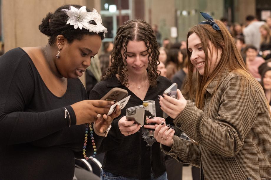 Three women huddle in a semi-circle with their phones in hand.