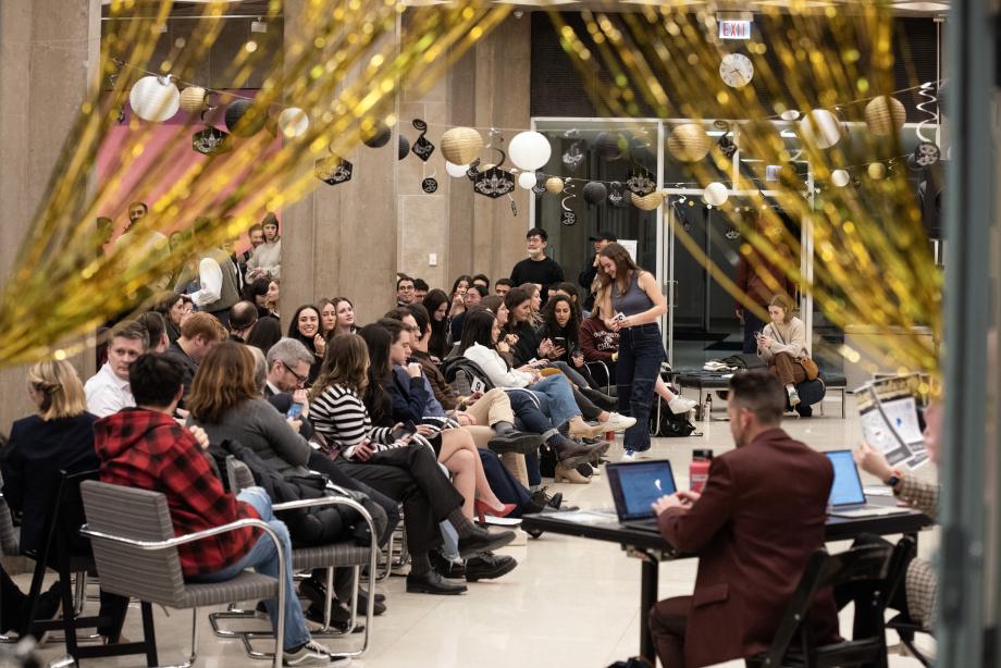 Students, faculty, and administrators sit in rows of chairs. Decor hangs from pillars and gold strands parted over a doorway frame the photo.