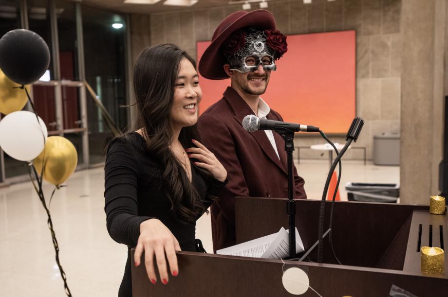 A woman stands at the podium in a black dress. A man in a maroon suit and metallic-looking masquerade mask looks on.