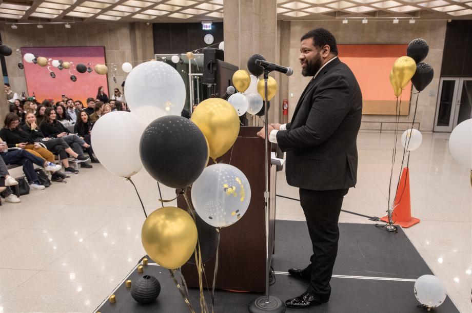 Adam Davidson stands at the podium surrounded by balloons. The audience is visible in the background.