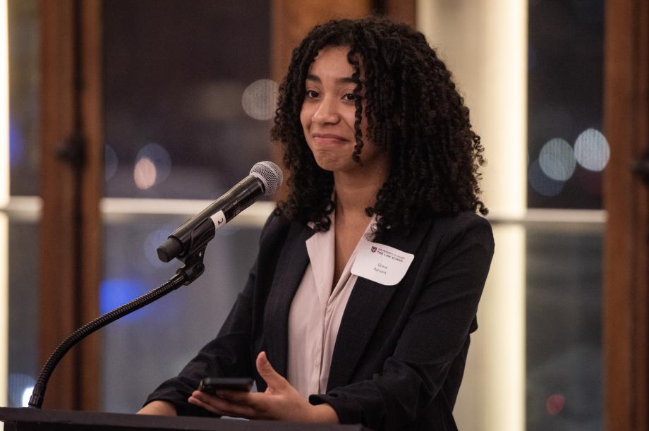 Grace Parsons wears a dark blazer over her blouse as she holds her phone and speaks into a microphone.