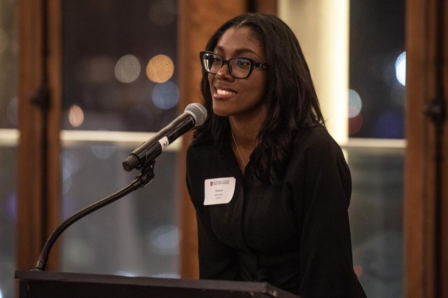 Briana Johnson wears a black blouse as she speaks into a microphone.