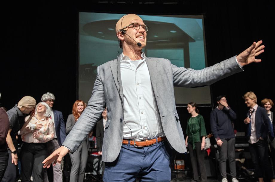 The student playing Professor Levmore, with a bald cap holds his arms outstretched as he sings.