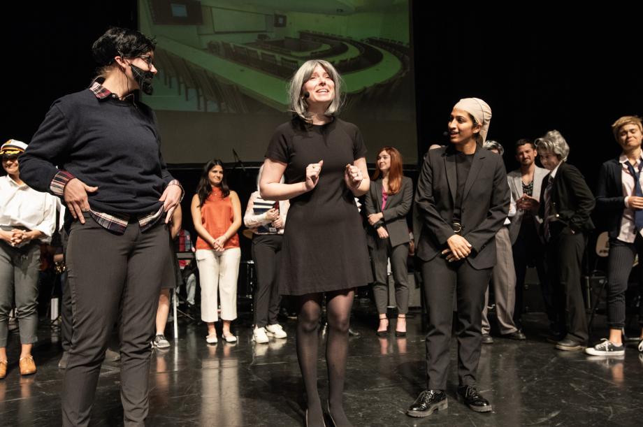 Three students in black and in wigs stand at the front of the stage.