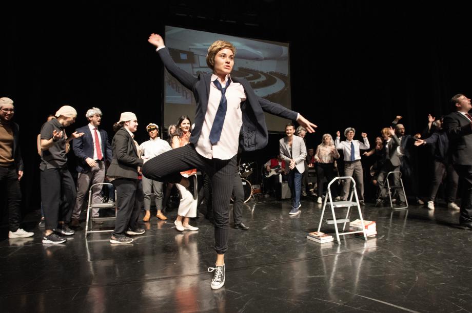A student in a suit and tie with a wig spins with one leg bent and arms outstretched.