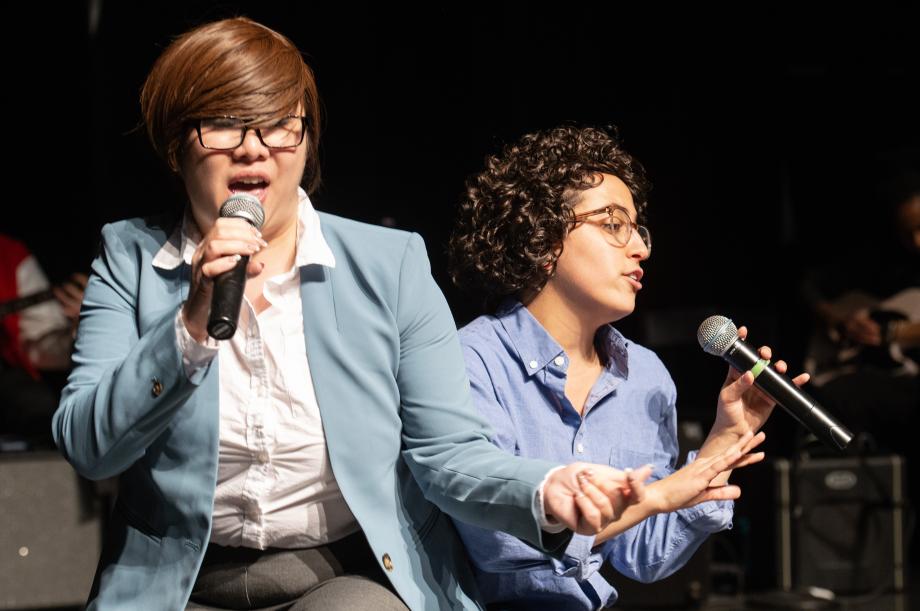 Two students in wigs and costumes hold microphones and dance.