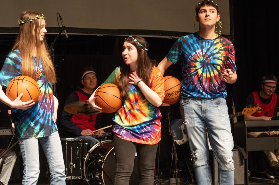 Three students with tie-dye shirts and flowers in their hair hold basketballs as they animatedly talk to each other.