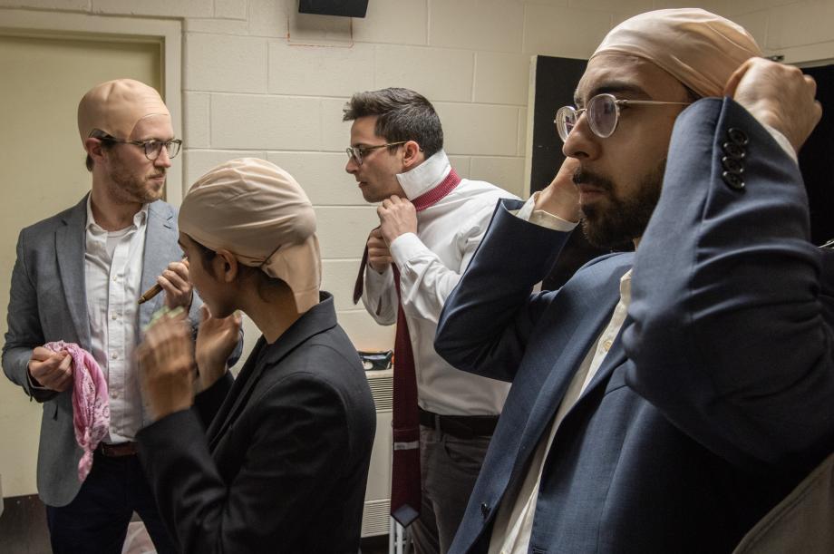Four students in a hallway adjust their wigs and bald caps. One ties a necktie.