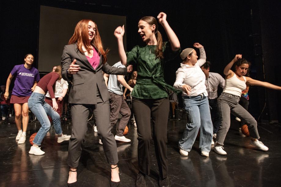 Students in wigs and costumes bump hips as they dance.
