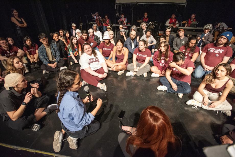 A couple dozen students sit in a semi-circle on the stag as one woman with a microphone provides direction.