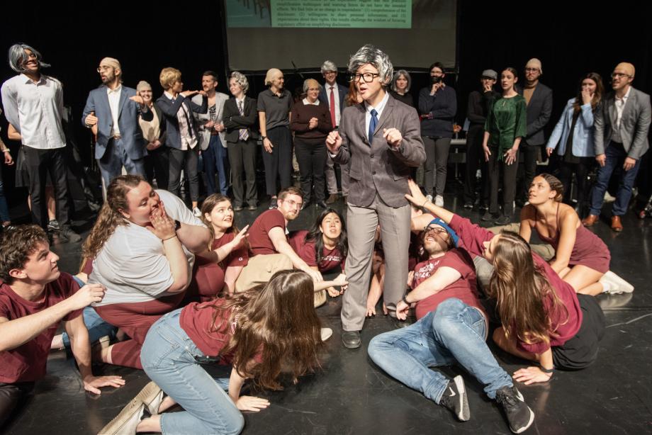 A student in costume and wig stands in the middle of a half-dozen students on the ground whose arms are reaching out to her.