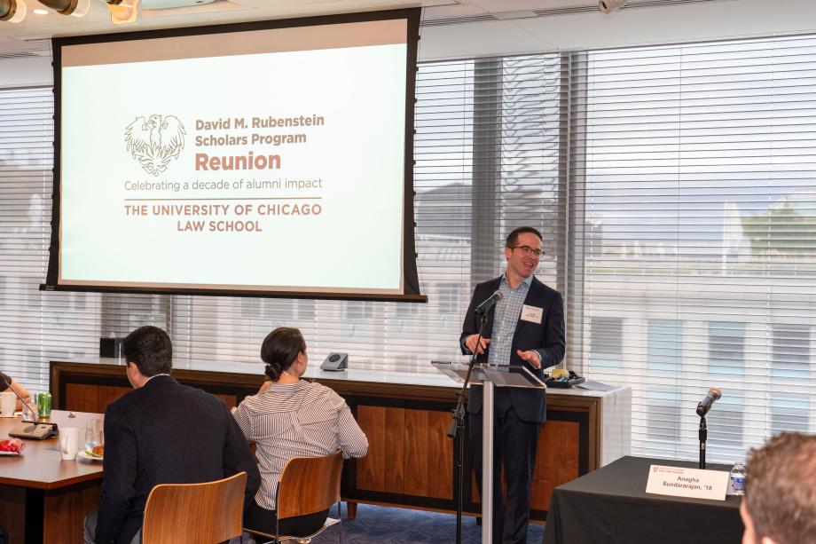 The Rubenstein Scholars Reunion kicked off with a day-time conference at Kirkland & Ellis LLP’s Washington, DC, office. Joe Schroeder, ’15, a partner at Kirkland and a Rubenstein Scholar alum, welcomed event attendees. Joe Schroeder, wearing a blue suit and open collar checkered shirt, stands at a podium to introduce the event. The display behind him says, “David M. Rubenstein Scholars Program Reunion. Celebrating a decade of alumni impact. The University of Chicago law School.