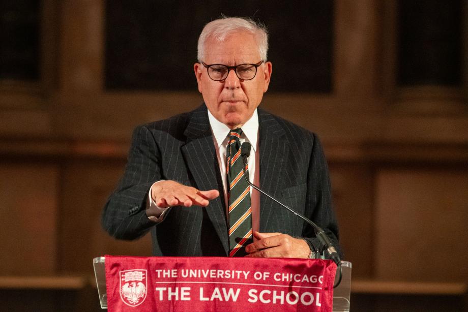 David M. Rubenstein spoke about why he created the Rubenstein Scholars Program and explained his involvement with the National Archives. David Rubenstein, wearing a pinstripe suit and striped tie, speaks at a podium draped with a University of Chicago Law School flag. He gestures, with his palm facing downward.