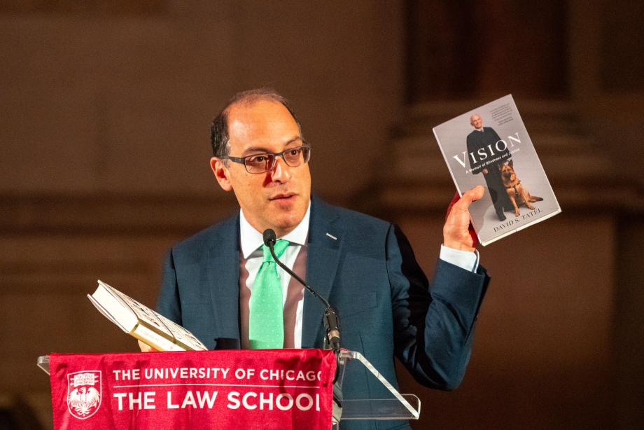 Professor Lior Strahilevitz introduced the Hon. David S. Tatel, ’66, and Tatel’s memoir, Vision. Lior Strahilevitz standing at a podium draped with a flag showing the UChicago Law logo. Strahilevitz speaks into the microphone while holding up a copy of Judge David Tatel's book, Visions, featuring David Tatel on the cover with his service animal.