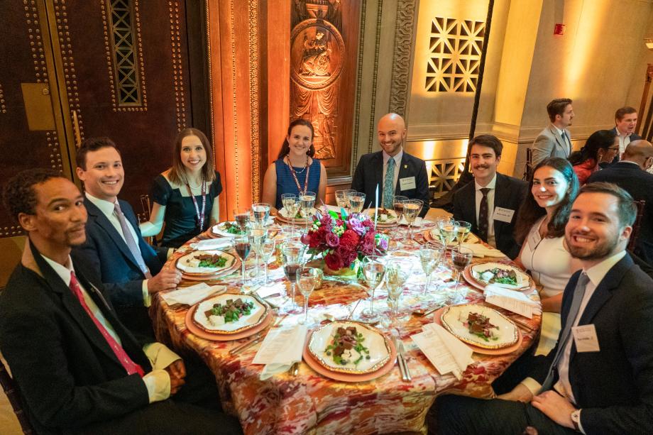 Rubenstein Scholars—graduates and current students alike—sat together over dinner. Event attendees sit around a circular table with plates of salad in front of them. All of the attendees at the table smile at the camera.