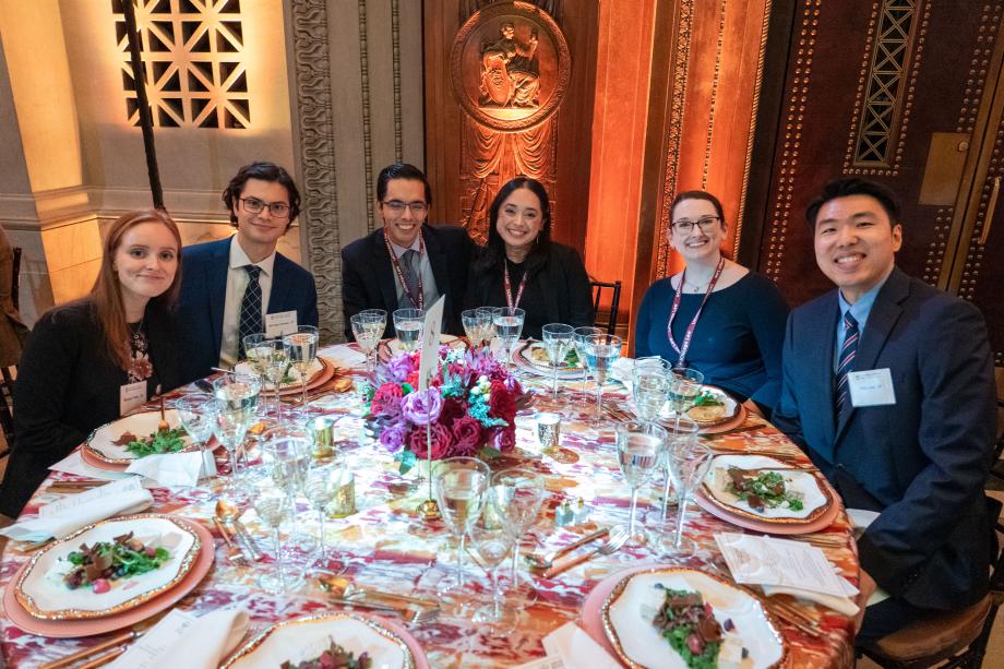 Dinner was held in the Rotunda for the Charters of Freedom in the National Archives Museum. The Rotunda is the home of America’s most treasured documents—the Declaration of Independence, the Constitution of the United States, and the Bill of Rights. Event attendees sit around a circular table with plates of salad in front of them. All of the attendees at the table smile at the camera.