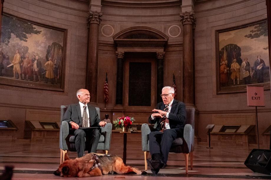 Tatel and Rubenstein discussed Vision during a fireside chat. Judge David Tatel sits with Rubenstein in individual chairs in front of the audience of attendees. Tatel smiles at Rubenstein while Rubenstein speaks, and Tatel's service animal lays on the floor in front of him.