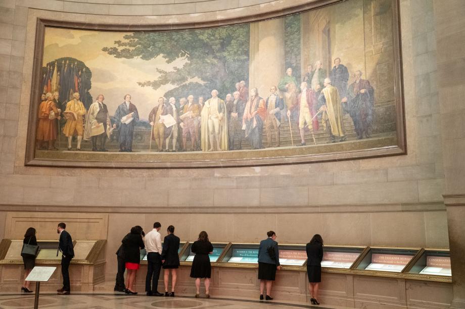 An evening dinner was hosted by David Rubenstein, ’73, at the National Archives Museum to celebrate the origins, goals, and transformative impact of the Rubenstein Scholars Program and provide another opportunity for Scholars to reminisce and reconnect with classmates and other Scholars. An enormous realistic painting of Revolutionary era American men is on the wall. Below the painting, a group of people look at documents behind glass cases.