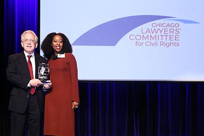 In a suit and tie, Jonathan Baum stands on a stage with a woman in a red dress. The screen behind them reads, "Chicago Lawyer's Committee for Civil Rights"