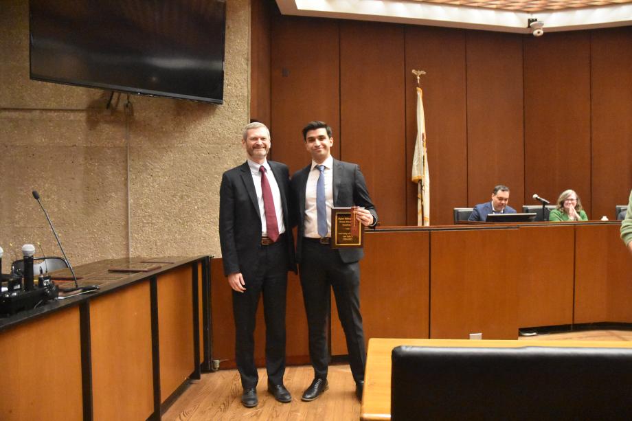 Two men wearing suits stand side-by-side posing together in a mock court room with one of the men holding a plaque.