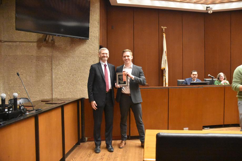 Two men wearing suits stand side-by-side posing together in a mock court room with one of the men holding a plaque.