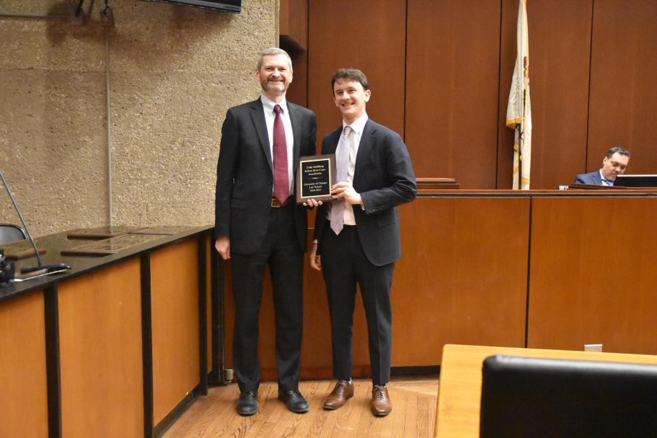Two men wearing suits stand side-by-side posing together in a mock court room with one of the men holding a plaque.