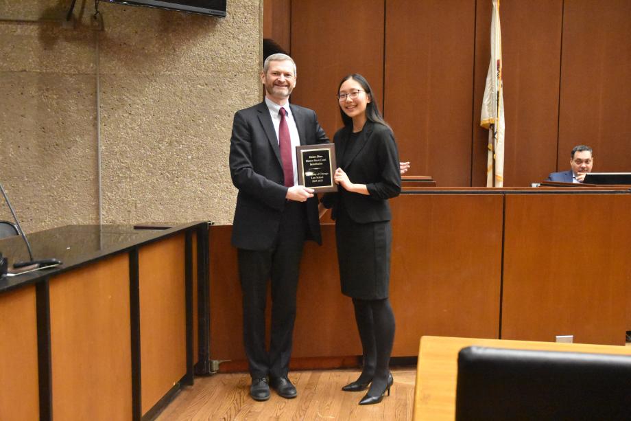 A man and a woman stand side-by-side posing together in a mock court room with the woman holding holding a plaque.