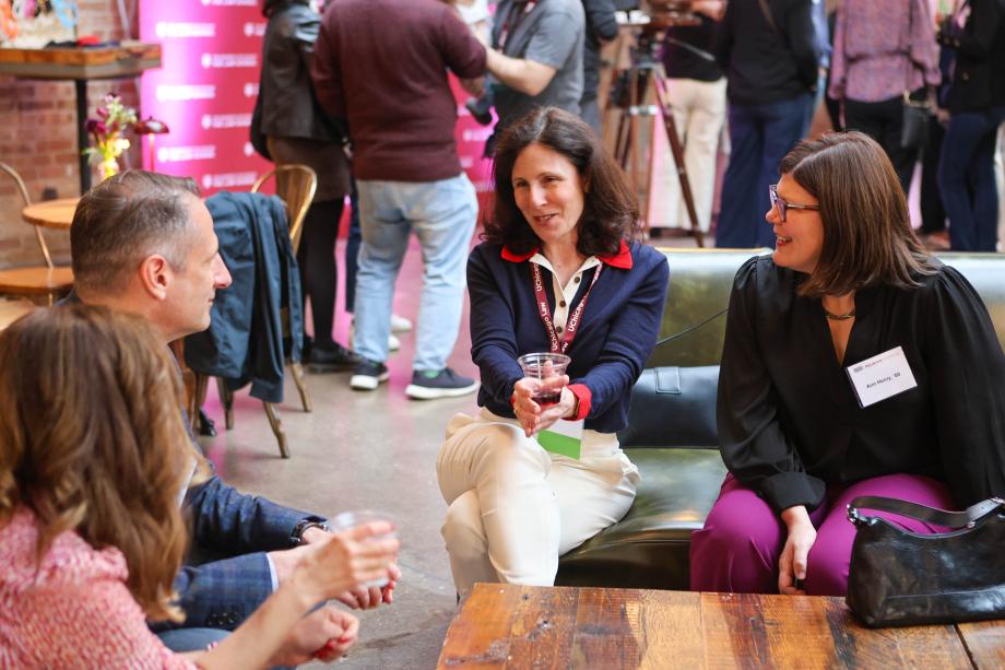 A group of people are sitting on couches talking to each other in a large open space filled with other people who are standing around and mingling.