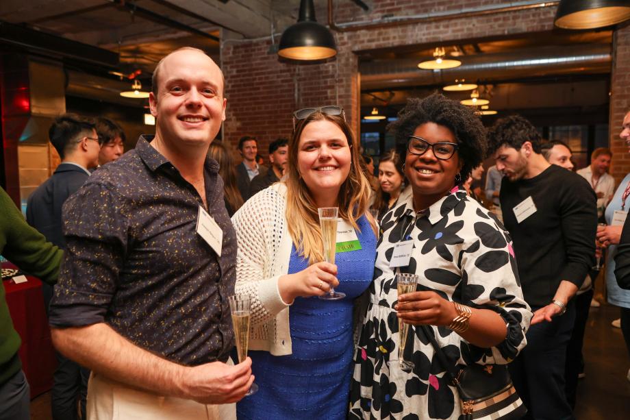 Three people are standing together smiling and posing for the camera while holding champagne glasses. The room is filled with people standing and mingling.