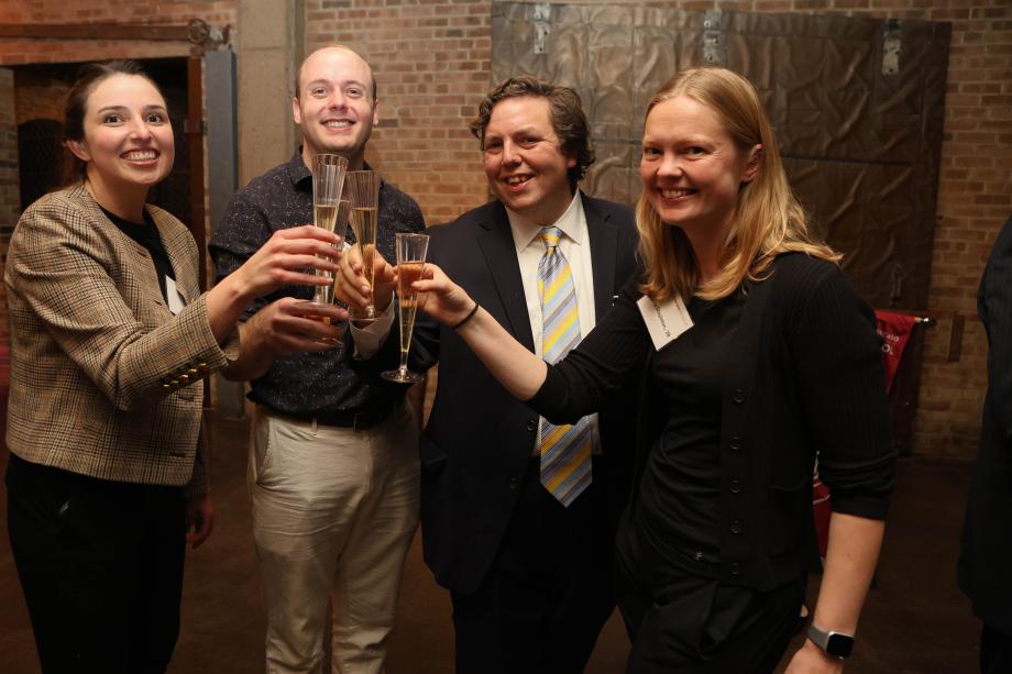 Four people are standing together smiling and offering cheers with champagne glasses. Behind them is a brick wall.