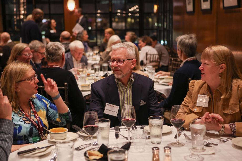 People are sitting and talking at a round dinner table in a busy restaurant. Dark windows at the back of the room indicate that it is evening time.