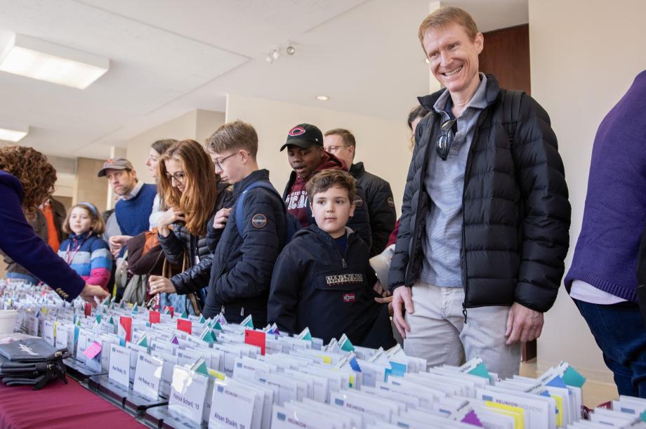 A man surrounded by children and other adults smiles down at a table filled with white name badges.