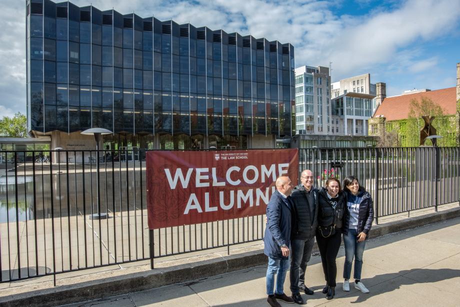 A view of the Law School building and blue skies with four people standing together in front of a banner that says Welcome Alumni.
