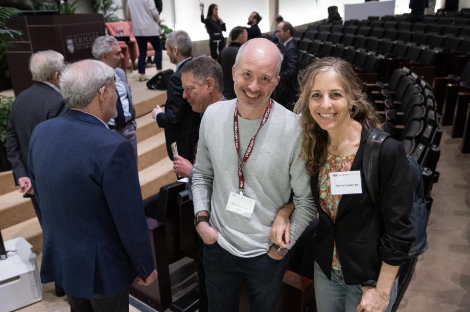 A man and a woman stand side by side smiling at the camera. They are in the auditorium that appears to be slowly emptying of people. A few audience members are standing around behind them.