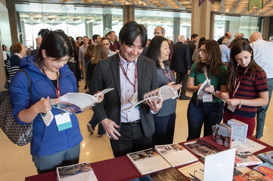  A man and a woman are looking through magazines while standing over a table located in a large open room filled with people