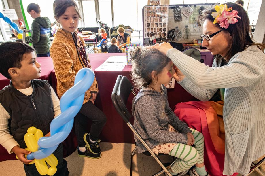 A woman is shown sitting on a chair and applying face paint to a child who is sitting directly in front of her. Other children are standing in line waiting to be face painted; one child is holding a blue balloon shaped into a spiral.